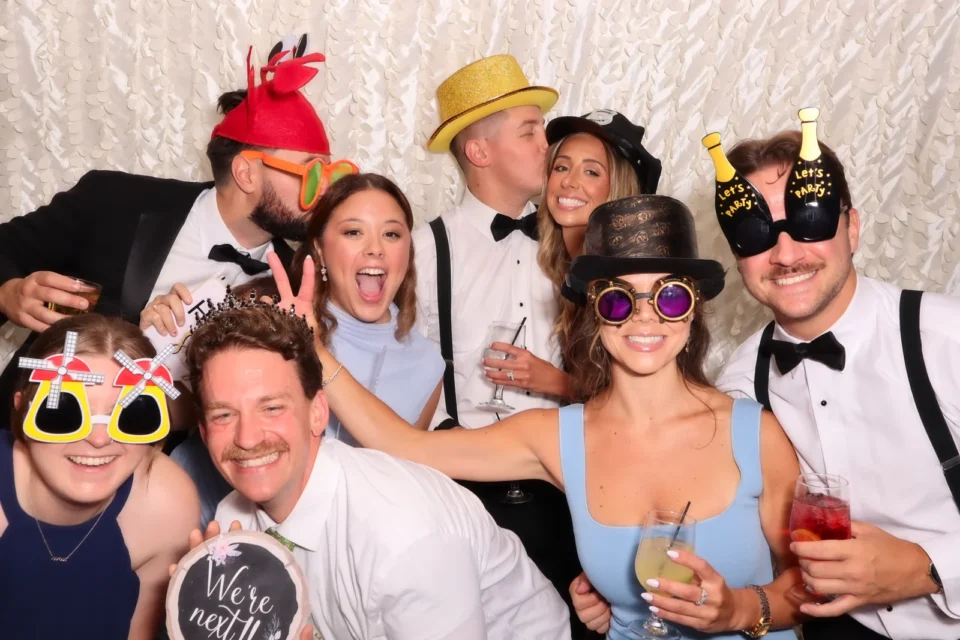 Open air photo booth rental NYC — wedding guests in hats and novelty glasses at a white backdrop