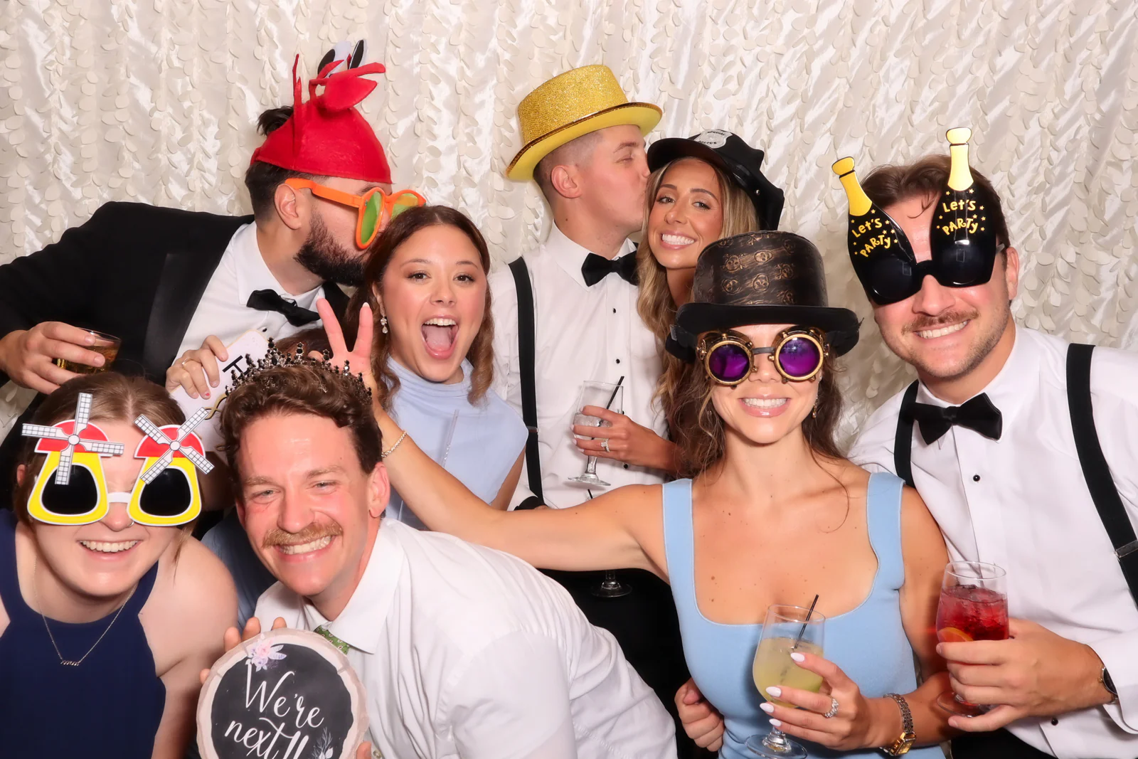 Open air photo booth rental NYC — wedding guests in hats and novelty glasses at a white backdrop